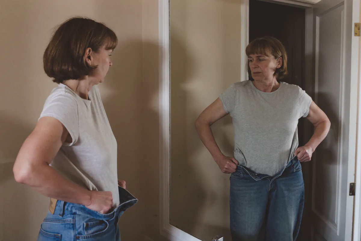 Frau vor dem Spiegel, die zu weite Jeans am Bund von sich wegzieht